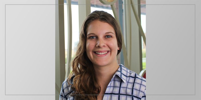 A white woman smiling with brown, wavy hair, wearing a blue and white patterned shirt.