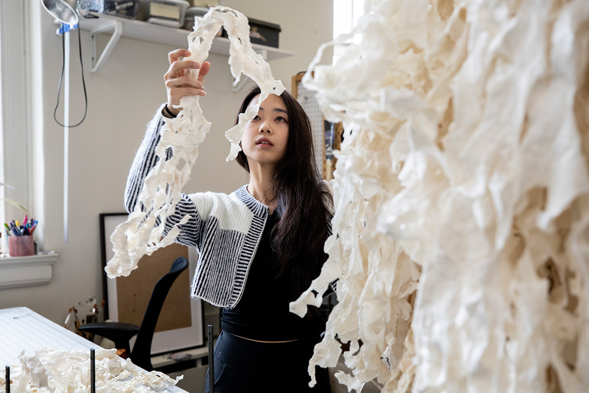Esther holds up and examines a large piece of wavy, off-white paper in her sunlit studio.