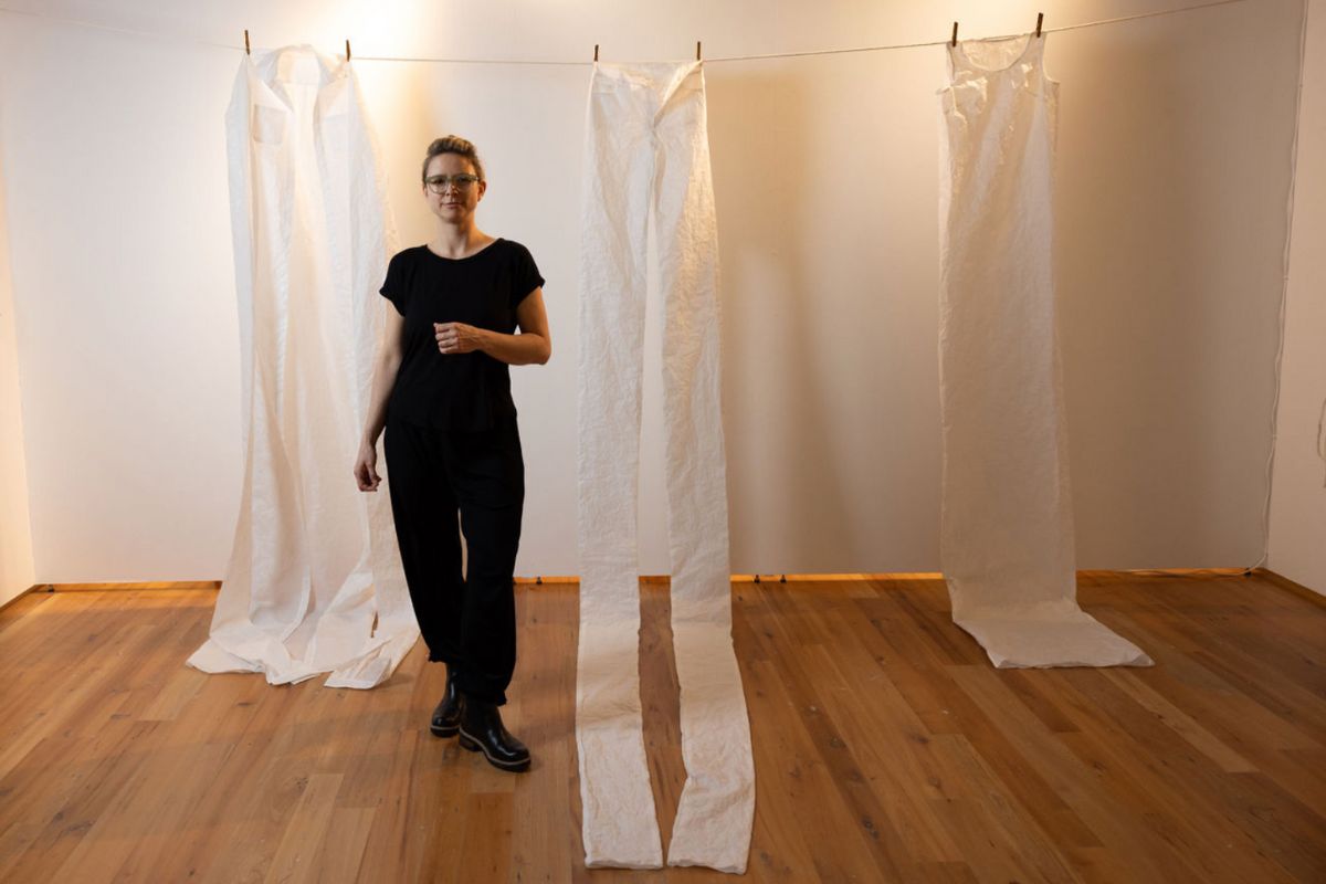 Heather stands in a gallery space and poses next to an oversized, white pair of pants and shirts hanging from a clothesline.