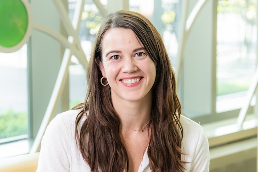 A white woman smiling, with brown, wavy hair, wearing a white shirt and gold earrings.
