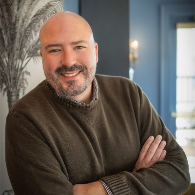A white male, bald, wearing a brown sweater with his arms crossed.