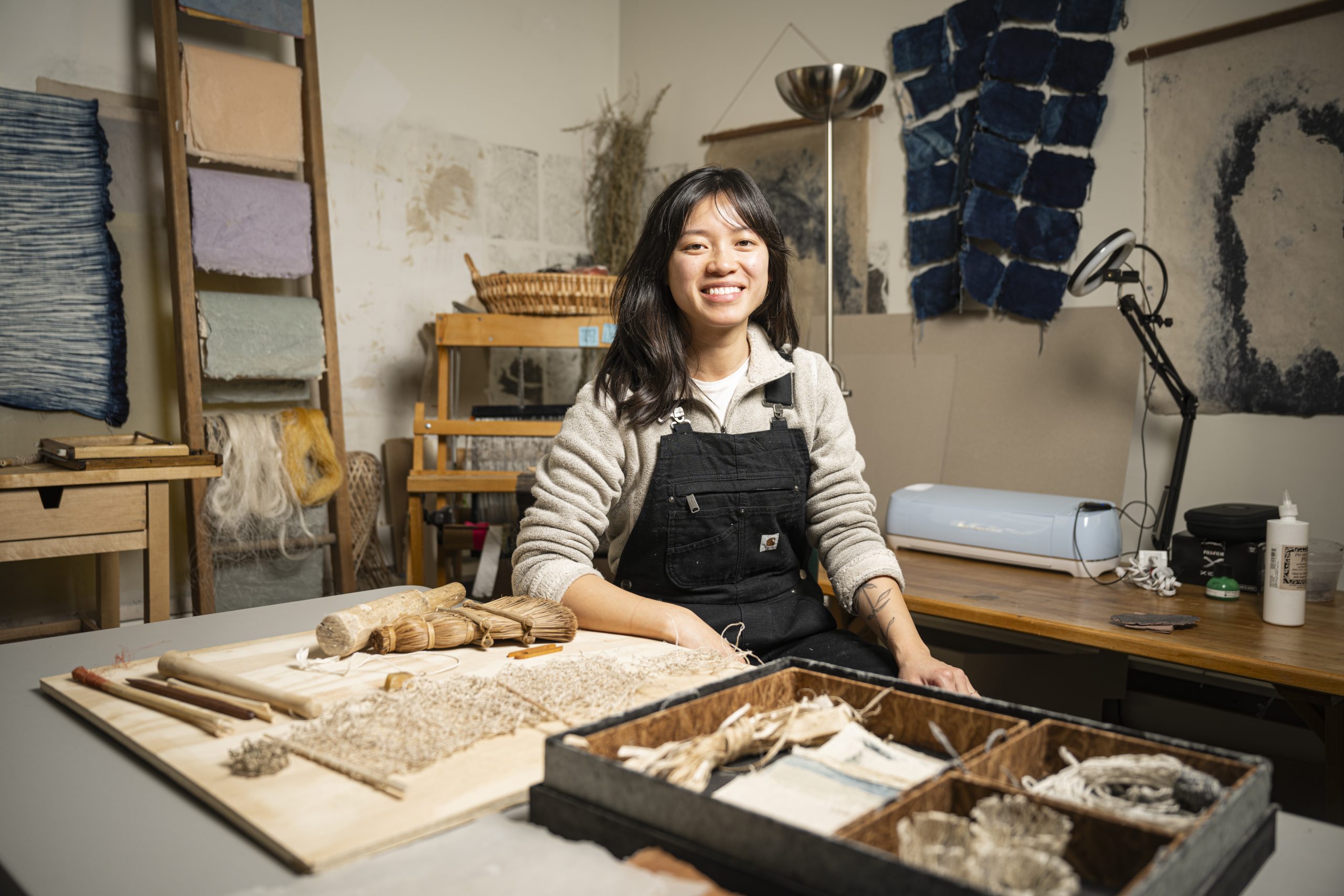 Veronica sits behind a table in the center, smiling and surrounded by equipment and artwork on the walls.