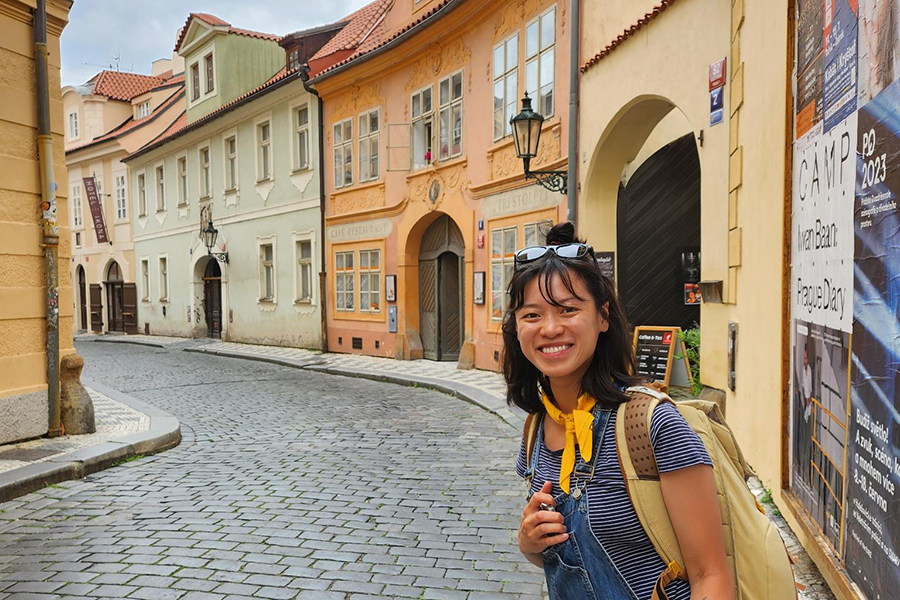 An Asian American woman with short, black hair poses and smiles on a street in Prague with pastel-colored buildings.