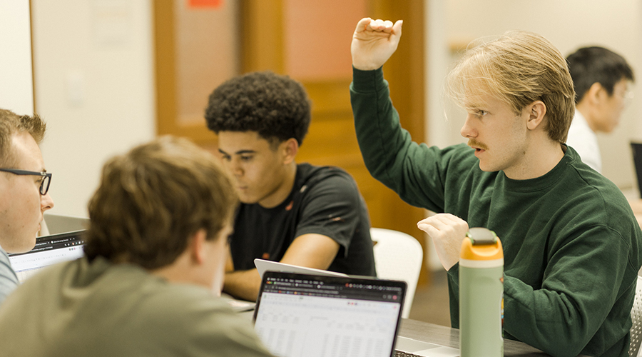 Students working at tables in small groups. One student gestures with their hands in the discussion.