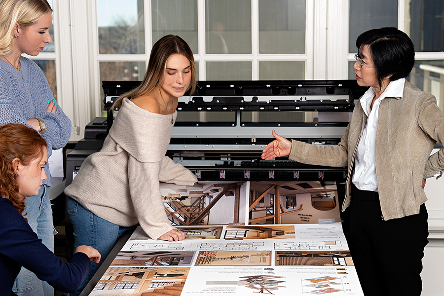 A professor speaks to three students as they look at a large format print.