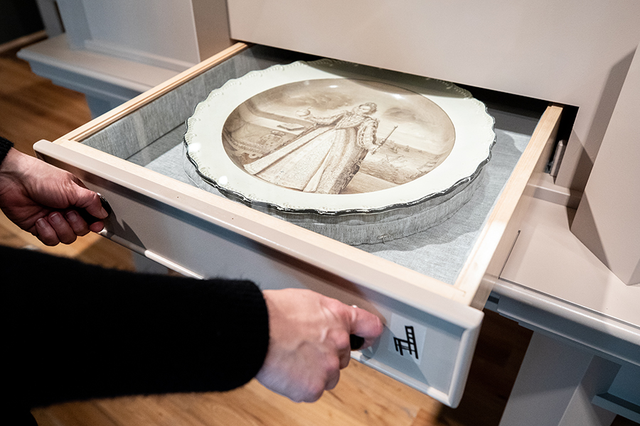 A hand opens a drawer displaying a plate under glass. The plate pictures Queen Elizabeth I in brown color tones.