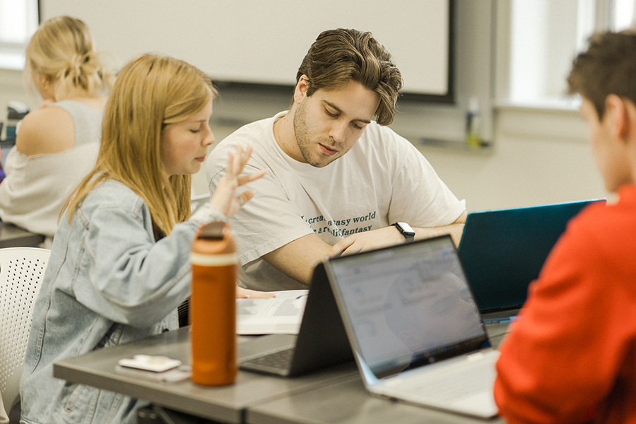 Students working at tables in small groups. Two students look down together at a paper, one is speaking and gesturing.