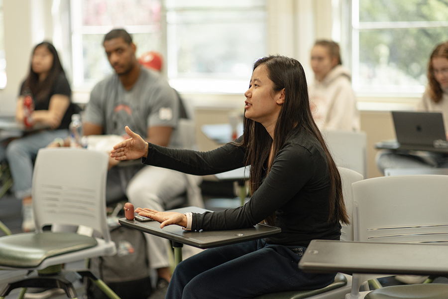 A student sits in a classroom, engaging in a discussion with their hand raised and surrounded by other students seated at desks.