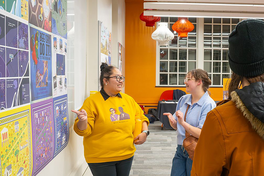 Two people stand near a wall of colorful posters in a brightly lit room. One person, wearing a yellow sweatshirt, gestures toward the posters while speaking. The other person, in a blue top and glasses, listens and smiles. A third person stands nearby with their back facing the camera.