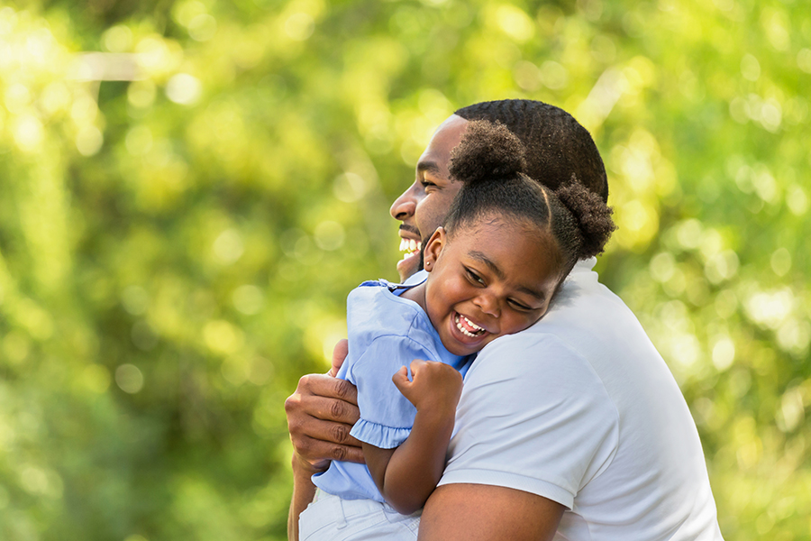 A black man smiles joyfully while hugging a black young girl who is also smiling and laughing. They are both outdoors with a background of green, blurry foliage on a sunny day. The girl wears a light blue dress and has her hair styled in two puffs.