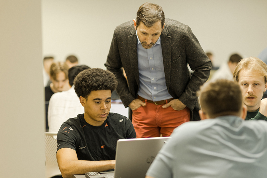 An instructor, stands and leans over to look at a laptop screen being used by a student sitting at a table with two other people engaged in conversation. The room is filled with people working.