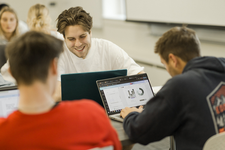 Three students seated at a table, collaboratively working on laptops. Other students work in the background.
