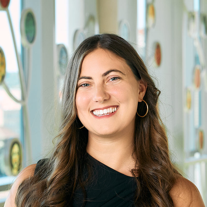 A white woman smiling with medium-length wavy brown hair, wearing a black dress, hoop earrings, and a septum ring.