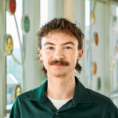 A white man smiling with a brown mullet and emerald tone button up shirt.