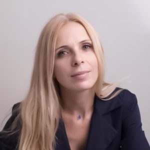 A white woman smiling, with long, blonde hair and blue eyes, wearing a navy blue suit.
