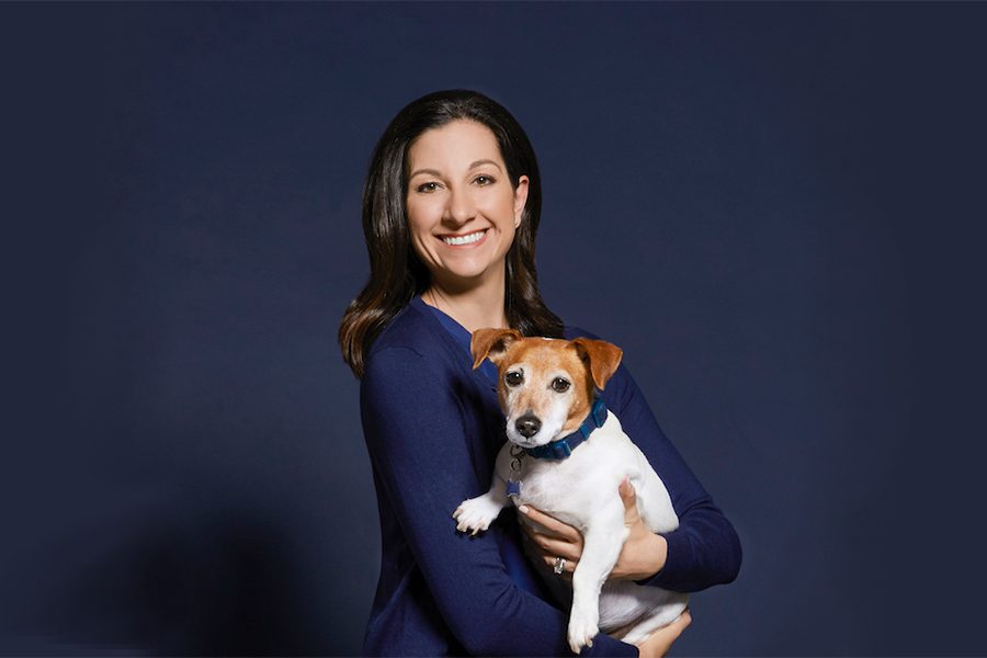 Francesca Mahoney poses in front of a navy blue background, in a navy blue shirt, holding her Jack Russell Terrier pet dog.