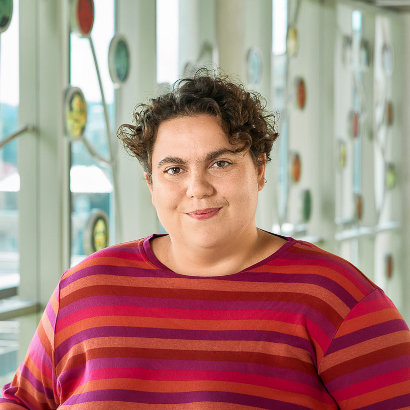 A white non-binary person with short, curly brown hair, smiling and wearing a bright striped tee shirt.