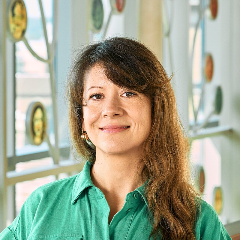 A white woman smiling, with long brown hair, wearing a collared green button-down shirt.