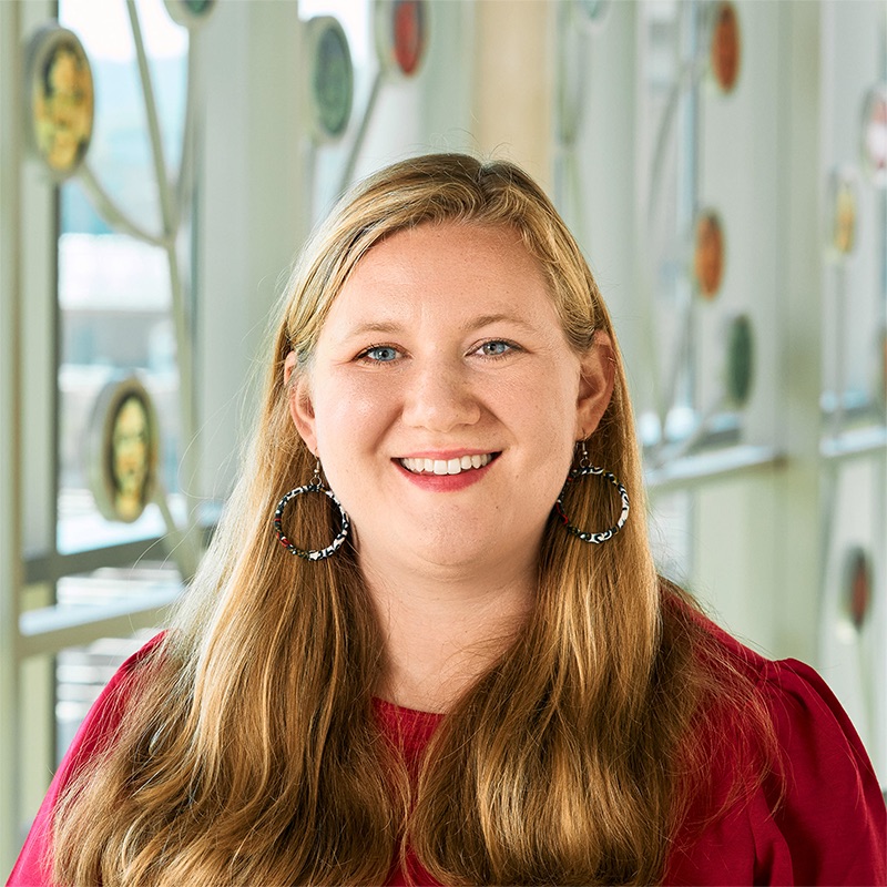 A white woman smiling, with long, straight blonde hair, wearing a red top and multi-colored hoop earrings.