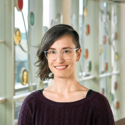 A white woman smiling with brown eyes and wavy brown hair that is shaved on one side, wearing clear plastic frame glasses and a purple sweater.