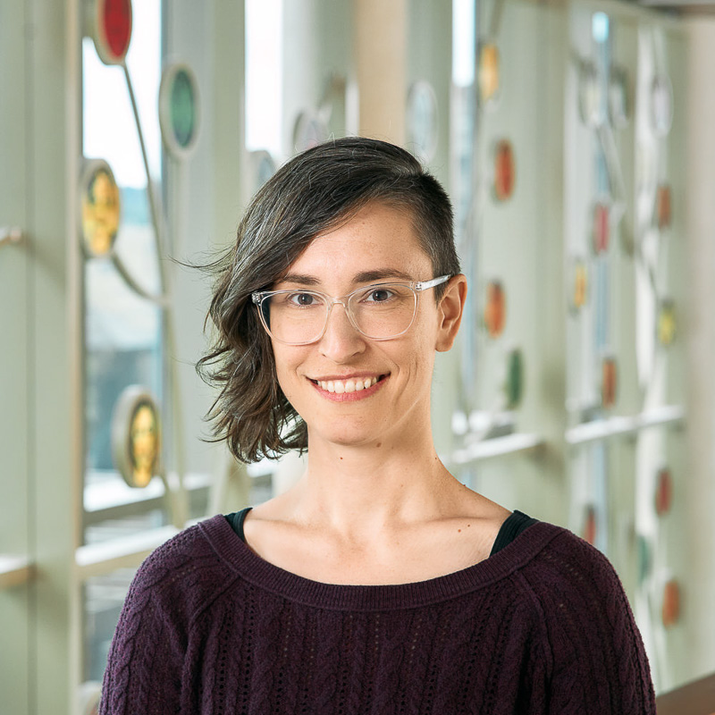 A white woman smiling with brown eyes and wavy brown hair that is shaved on one side, wearing clear plastic frame glasses and a purple sweater.
