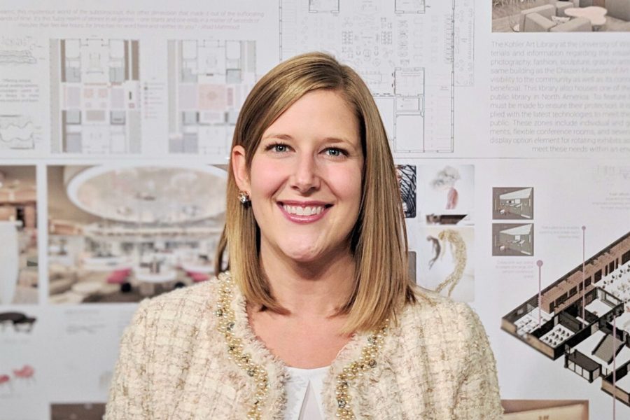 A woman with shoulder-length blonde hair smiles while standing in front of a wall that displays designs for an architecture project.