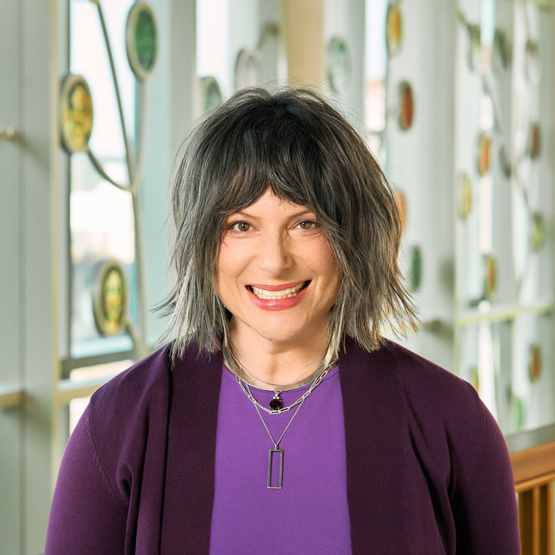 A white woman smiling, with shoulder length brown hair, wearing a dark plum sweater with a purple shirt and silver necklaces.