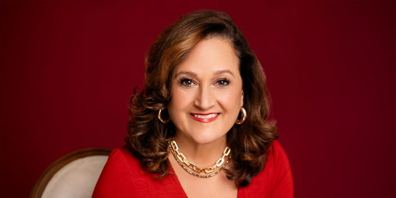 A professionally dressed woman smiling, wearing a red shirt on a deep red background.