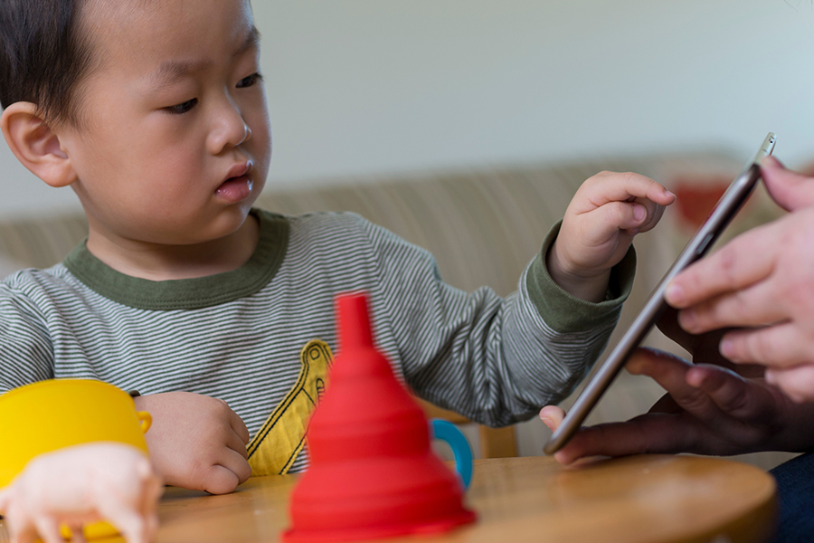 A small child at a table with toys points to a tablet held by an adult's hand.