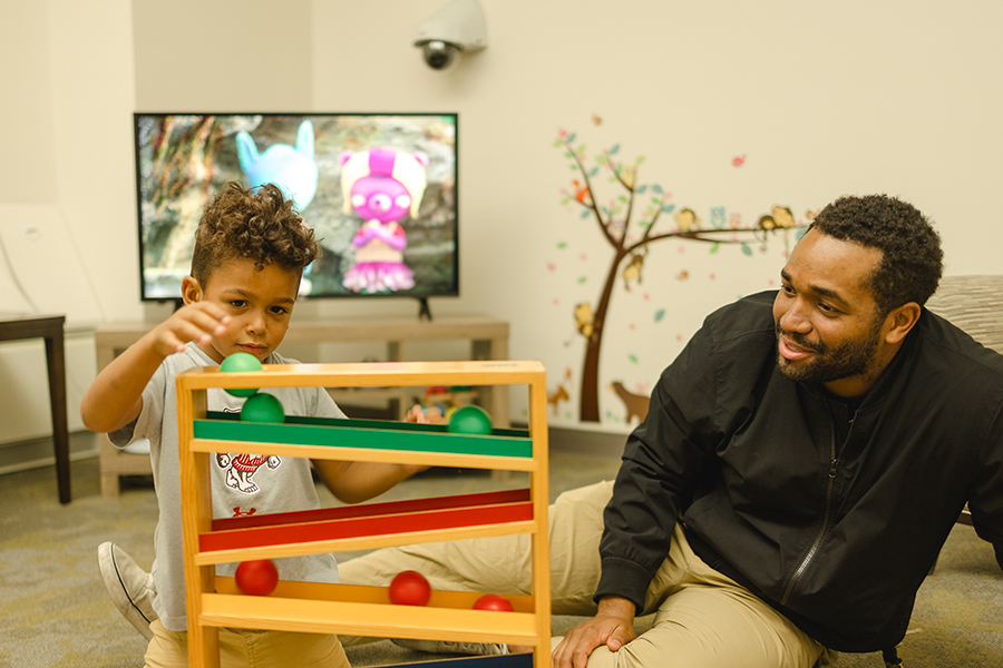 A child plays with a ball and ramp toy while their parent watches and smiles. They are in a research lab set as a living room space.