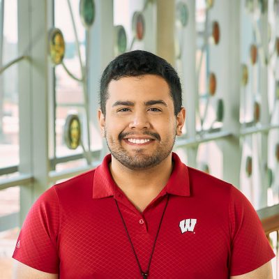 A brown man smiling with short black har, wearing a red shirt and necklace.