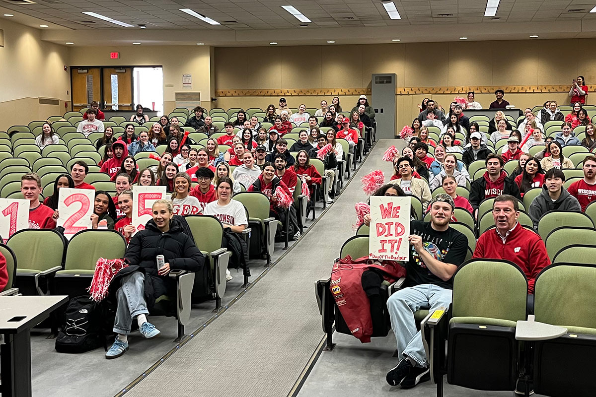 A lecture hall full of undergraduate students. Three students hold a sign that says "125", and one student holds a sign that says "We did it!".