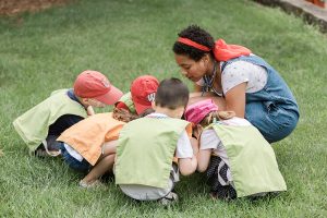 A student kneels in a group of children all looking at something in the grass.