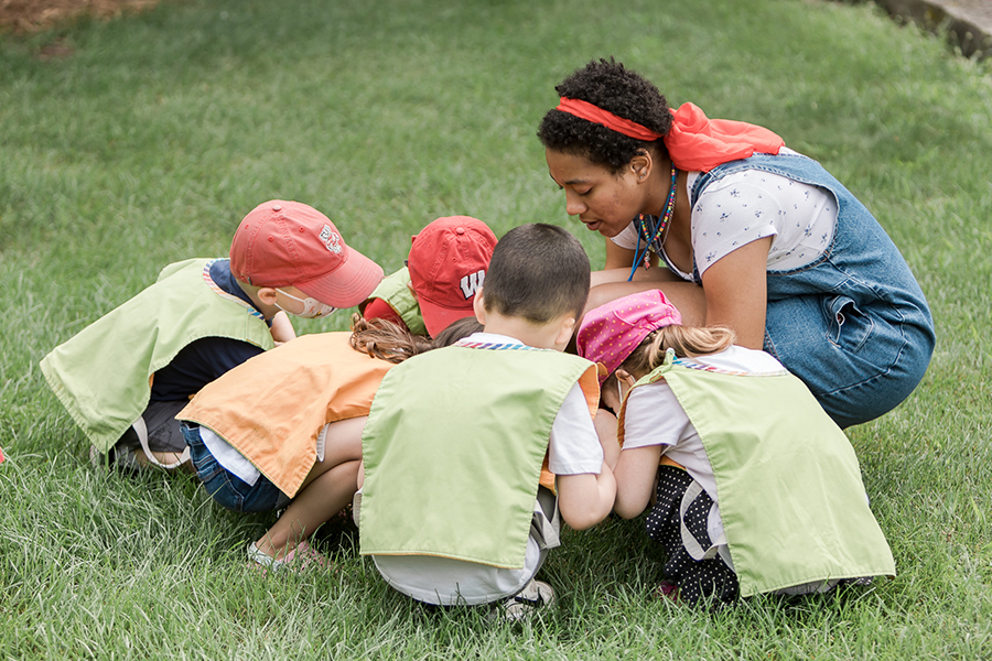 A student kneels in a group of children all looking at something in the grass.