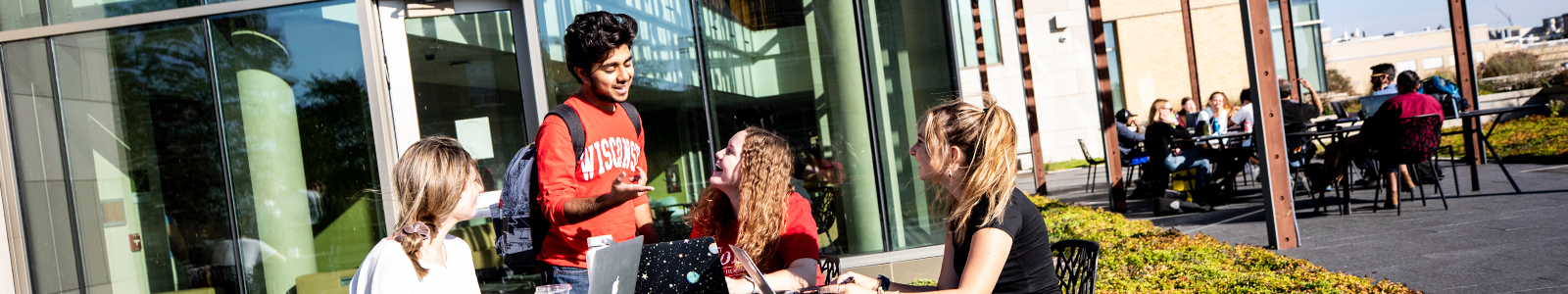 Students talking and studying around a table on the school rooftop terrace on a sunny day. A group of people sit at tables in the background.