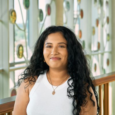 A brown woman smiling with long black curly hair, wearing a white shirt and a gold necklace.