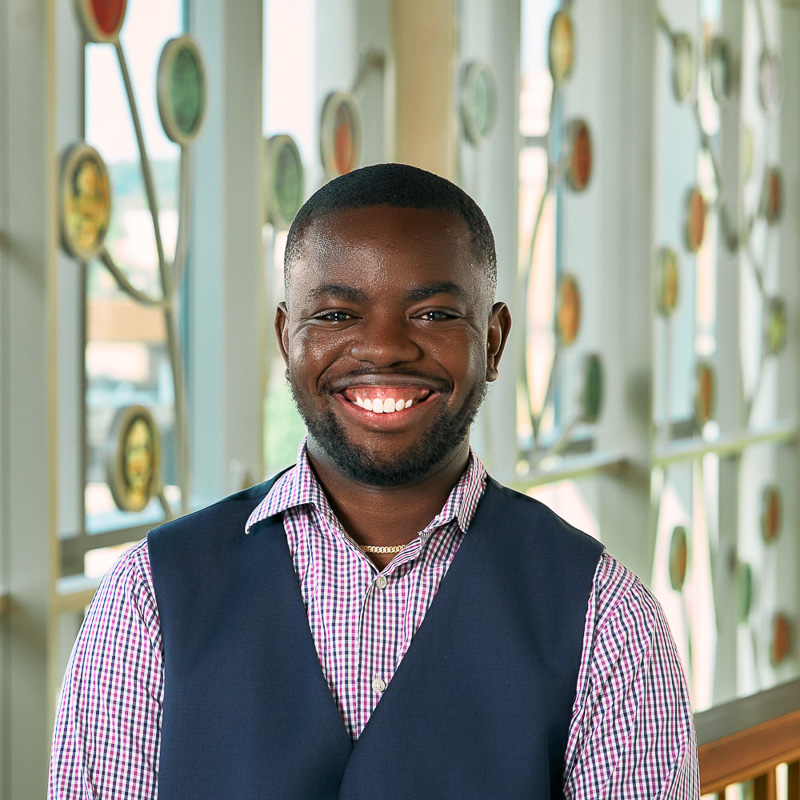 A Black man with a warm, wide smile and short-cropped black hair, wearing a blue, white and pink checkered button-down shirt layered under a navy blue vest, with a thin gold chain visible at his neck.