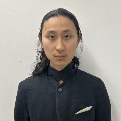 An East Asian male staring into the camera, with long black hair tied back, wearing a black linen jacket.