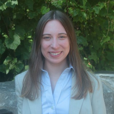A white woman smiling, with brown, straight hair, wearing a blue shirt and a brown blazer, posing in front of a tree.