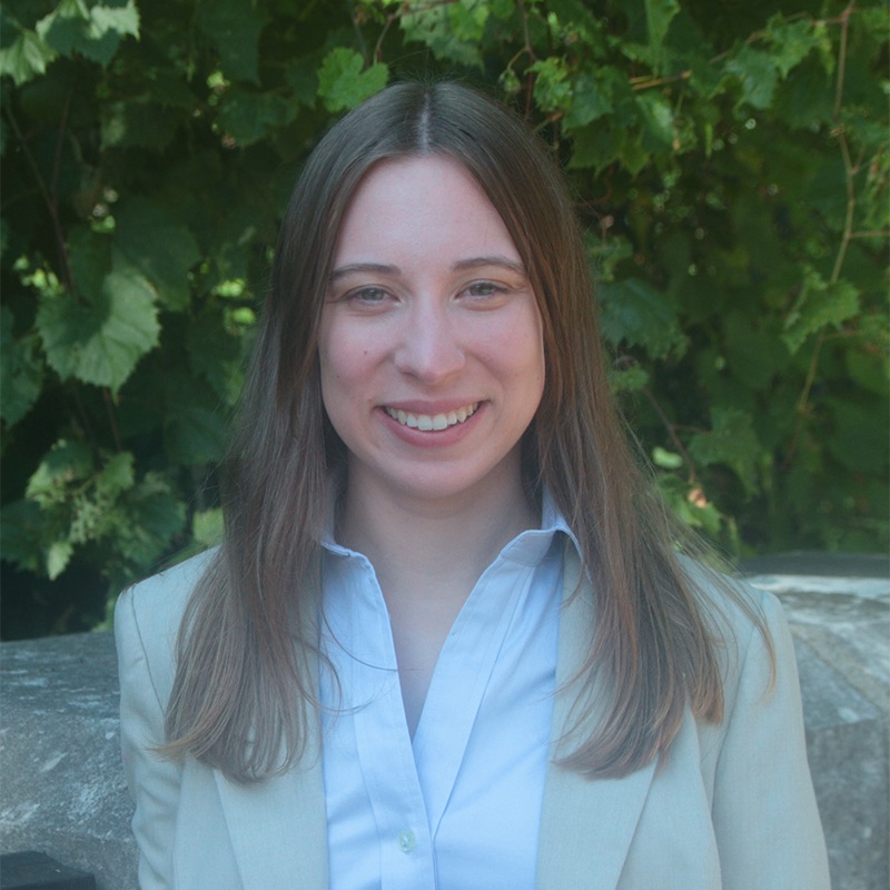 A white woman smiling, with brown, straight hair, wearing a blue shirt and a brown blazer, posing in front of a tree.