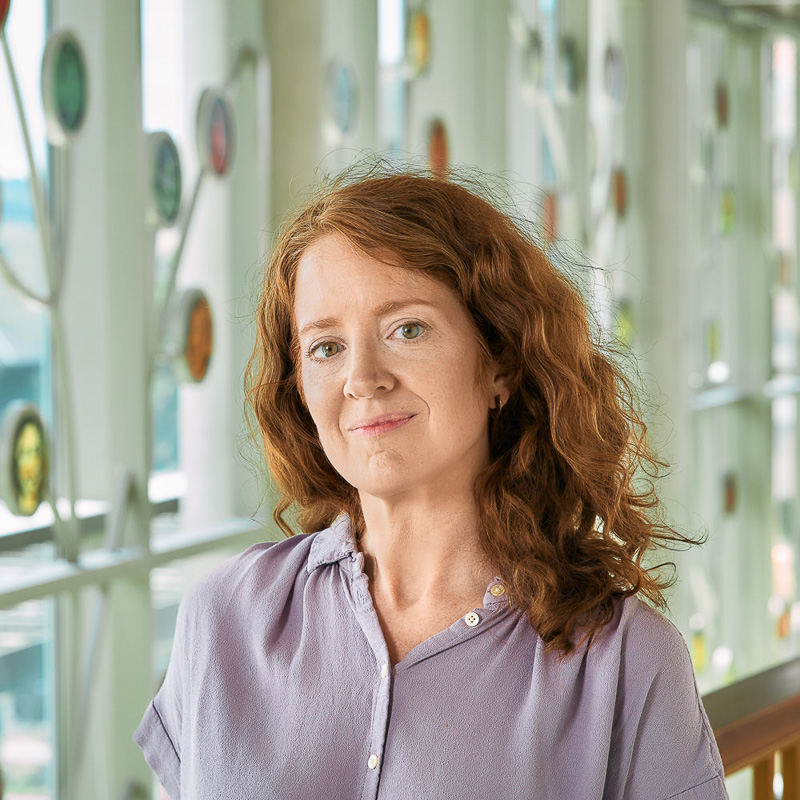 A white woman with red, curly hair, wearing a lavender shirt.