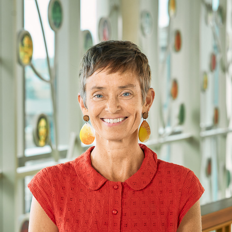 A white woman smiling, with short brown hair, wearing an orange top and yellow teardrop earrings.