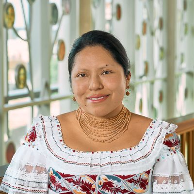 Indigenous woman smiling, with black long straight hair, wearing a white blouse with a gold necklace and earrings.