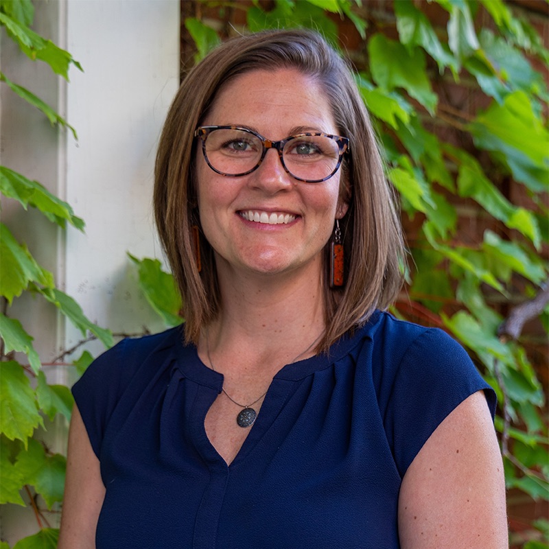 A white woman smiling, with brown hair, wearing glasses and a blue shirt, in front of an ivy and brick background.