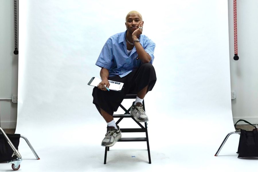 G Green sits on a stool in front of a white backdrop on a photography set, leaning forward, their elbow resting on their bent knee, hand supporting their chin. Their opposite hand holds a pen and clipboard. G has a content expression looking at the camera.