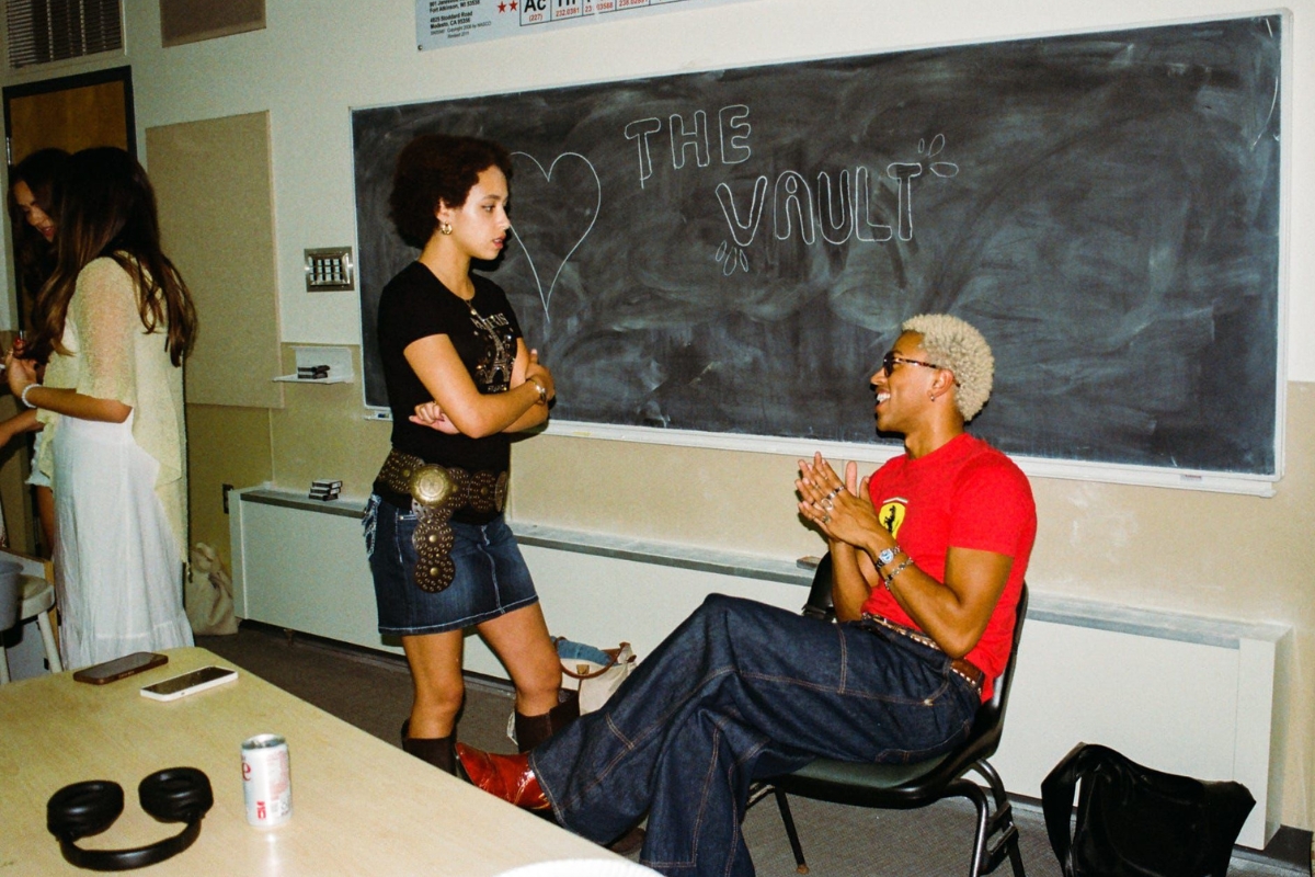 G sits in front of a chalkboard that says, "The Vault" with a big heart drawn next to it. G is smiling, talking with Philbert. Others are talking behind them.
