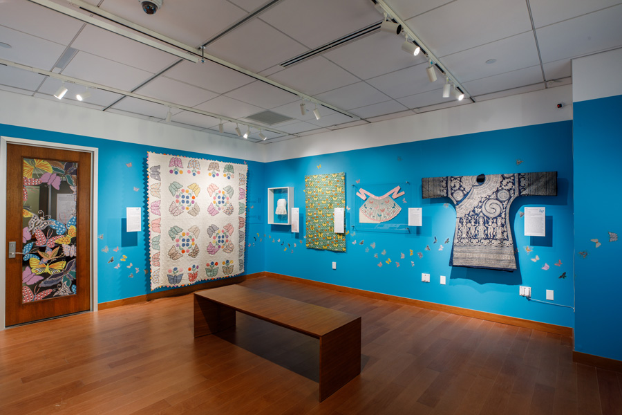 A collection of textiles on a blue wall with bench centered in the middle of the gallery.
