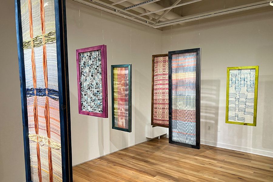 Six textiles of different shapes and colors, hang in long, rectangular frames from a gallery ceiling.