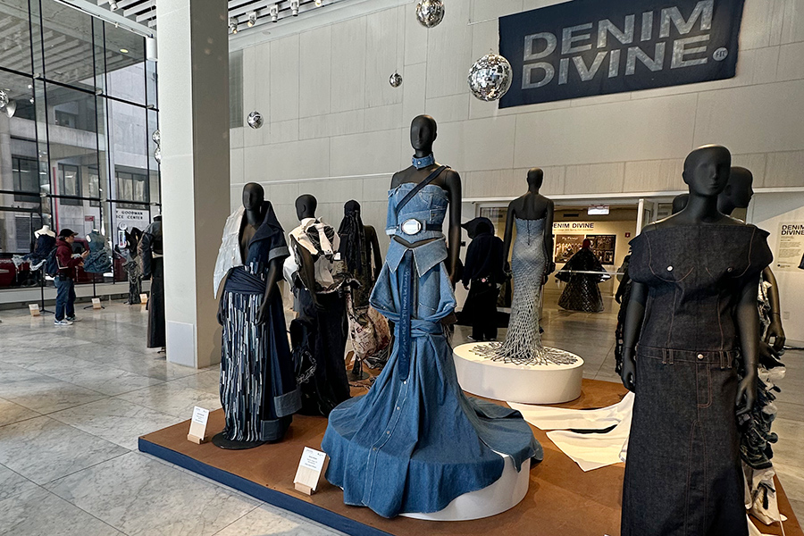 A display of mannequins with long gowns are in the large, tan brick entryway of a building with large, floor to ceiling windows. A sign on the wall in the background says, Denim Devine.
