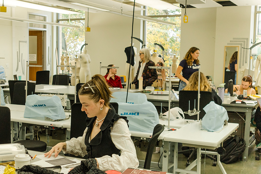Studio classroom of student drawing at large tables. A student and instructor talk in the background.